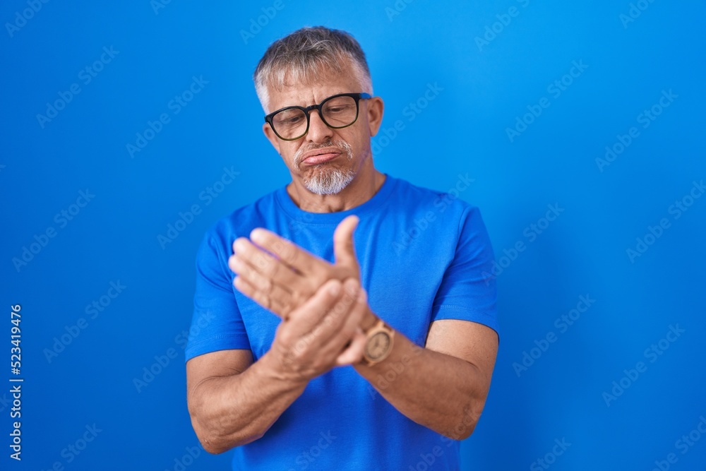 Hispanic man with grey hair standing over blue background suffering pain on hands and fingers, arthritis inflammation Hispanic man with grey hair standing over blue background suffering pain on hands and fingers, arthritis inflammation