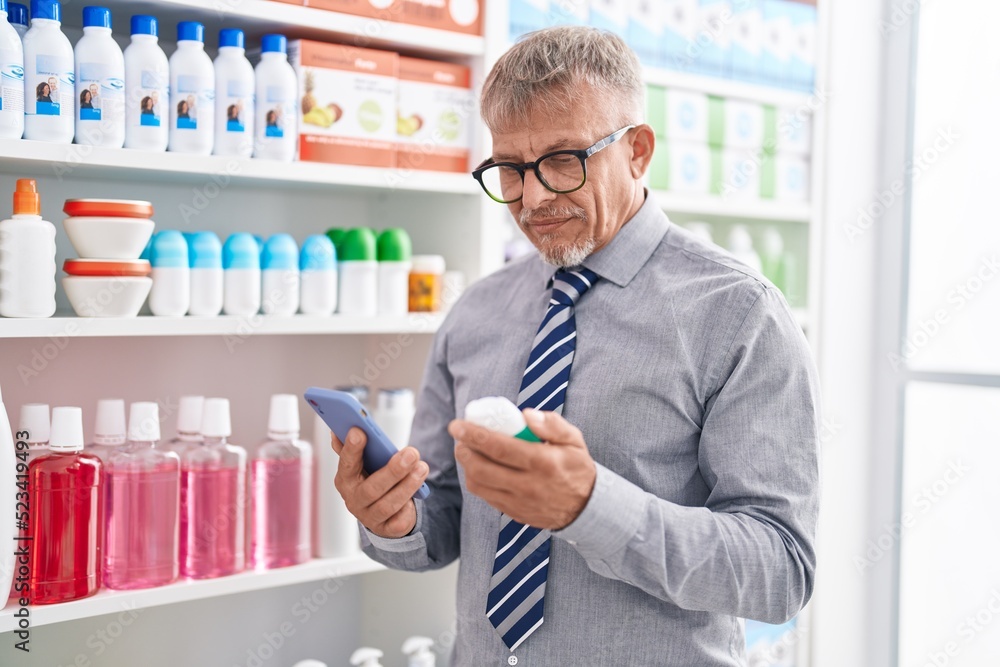 Middle age grey-haired man customer using smartphone holding medicine bottle at laboratory Middle age grey-haired man customer using smartphone holding medicine bottle at laboratory