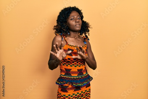 Young african american woman wearing traditional african clothes disgusted expression  displeased and fearful doing disgust face because aversion reaction. with hands raised