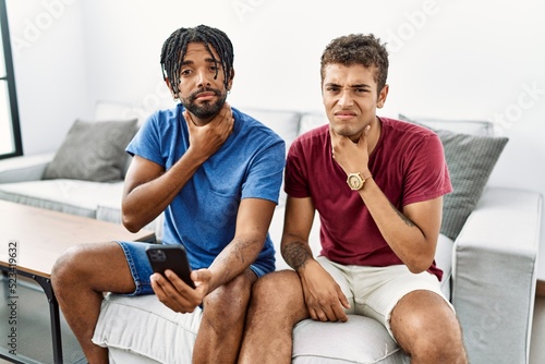 Young hispanic men using smartphone sitting on the sofa at home touching painful neck, sore throat for flu, clod and infection