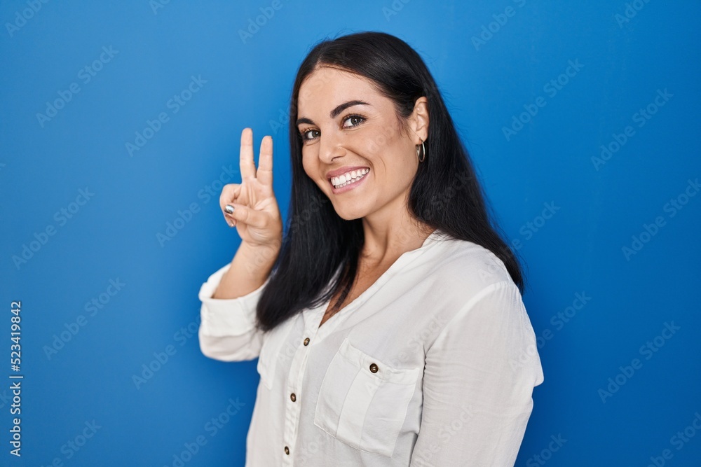 Young hispanic woman standing over blue background smiling looking to the camera showing fingers doing victory sign. number two. Young hispanic woman standing over blue background smiling looking to the camera showing fingers doing victory sign. number two.