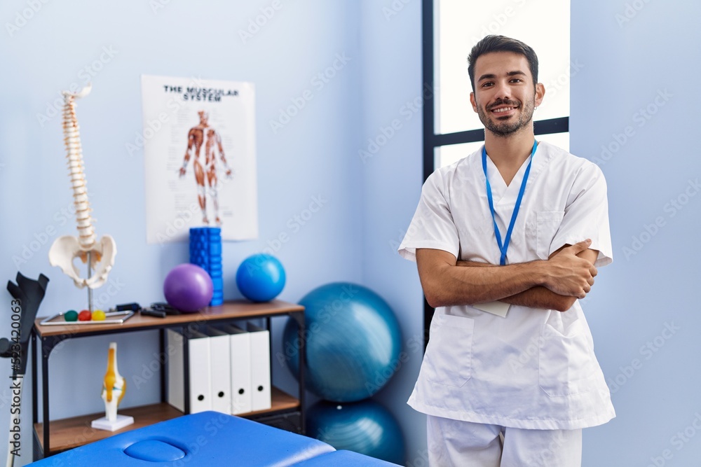 Young hispanic man wearing physiotherapist uniform standing with arms crossed gesture at rehab clinic - obrazy, fototapety, plakaty 