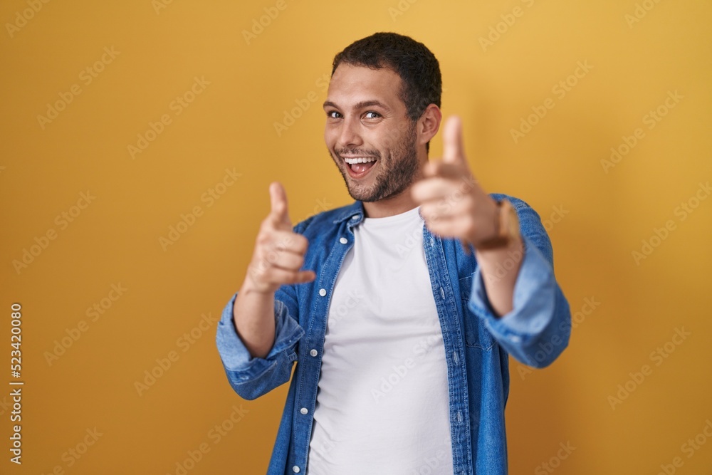 Hispanic man standing over yellow background pointing fingers to camera with happy and funny face. good energy and vibes. Hispanic man standing over yellow background pointing fingers to camera with happy and funny face. good energy and vibes.