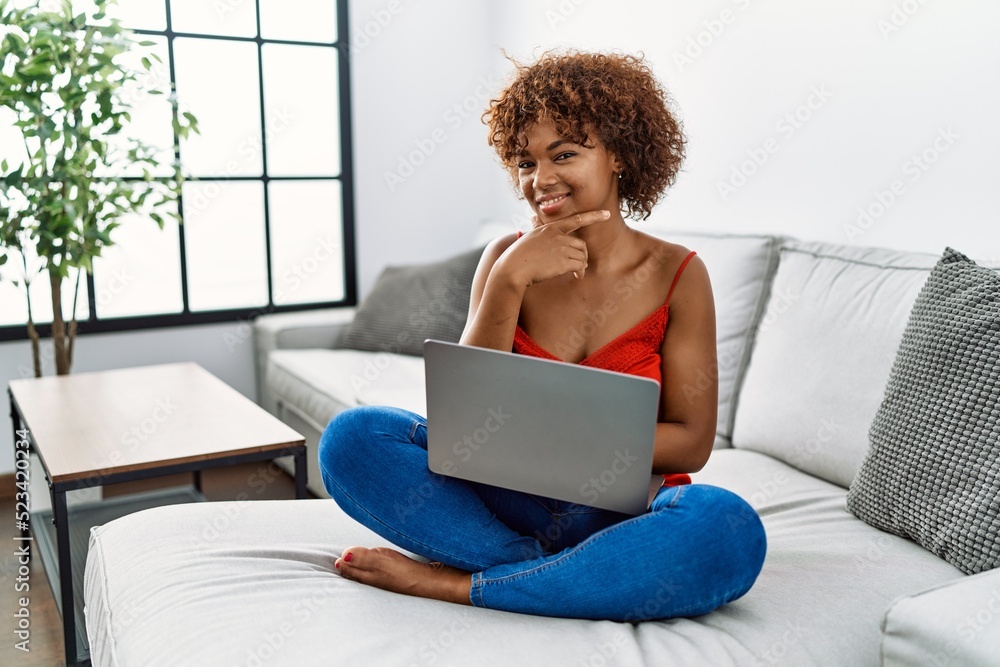 Young african american woman sitting on the sofa at home using laptop looking confident at the camera smiling with crossed arms and hand raised on chin. thinking positive. Young african american woman sitting on the sofa at home using laptop looking confident at the camera smiling with crossed arms and hand raised on chin. thinking positive.