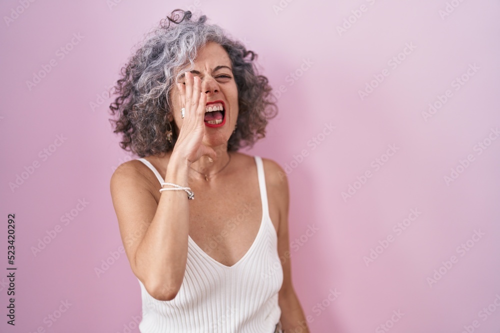 Middle age woman with grey hair standing over pink background shouting and screaming loud to side with hand on mouth. communication concept.