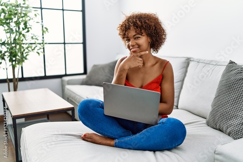 Young african american woman sitting on the sofa at home using laptop looking confident at the camera smiling with crossed arms and hand raised on chin. thinking positive.