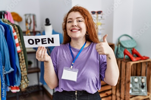 Young redhead woman holding banner with open text at retail shop smiling happy and positive  thumb up doing excellent and approval sign