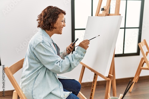 Middle age hispanic artist woman using smartphone drawing at art studio.