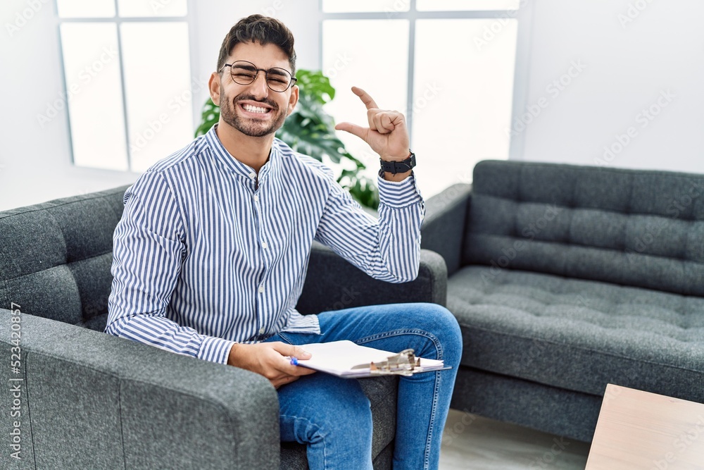 Young psychologist man at consultation office smiling and confident gesturing with hand doing small size sign with fingers looking and the camera. measure concept. Young psychologist man at consultation office smiling and confident gesturing with hand doing small size sign with fingers looking and the camera. measure concept.