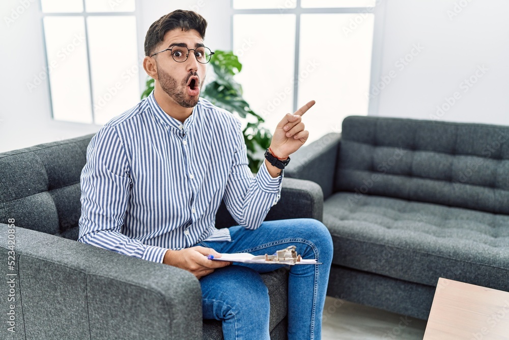 Young psychologist man at consultation office surprised pointing with finger to the side, open mouth amazed expression. Young psychologist man at consultation office surprised pointing with finger to the side, open mouth amazed expression.