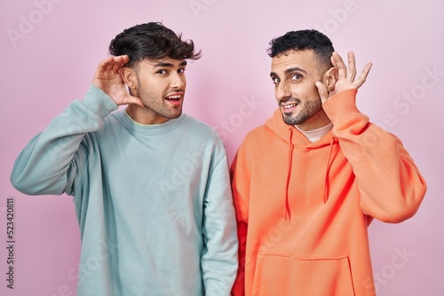 Young hispanic gay couple standing over pink background smiling with hand over ear listening an hearing to rumor or gossip. deafness concept.