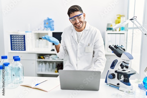 Young hispanic man working at scientist laboratory with laptop celebrating achievement with happy smile and winner expression with raised hand