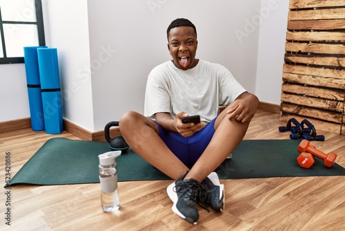 Young african man sitting on training mat at the gym using smartphone sticking tongue out happy with funny expression. emotion concept.