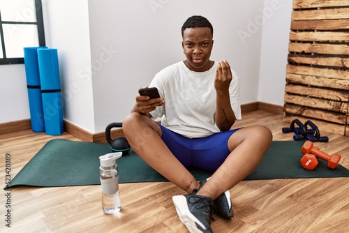 Young african man sitting on training mat at the gym using smartphone doing italian gesture with hand and fingers confident expression
