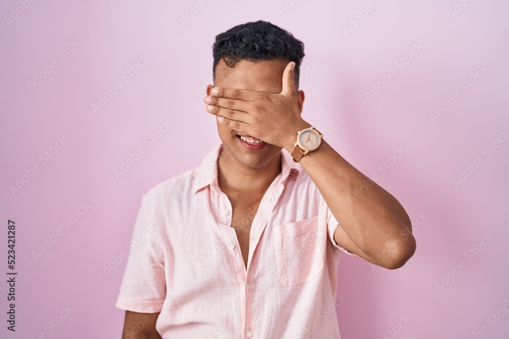 Young hispanic man standing over pink background covering eyes with hand, looking serious and sad. sightless, hiding and rejection concept Young hispanic man standing over pink background covering eyes with hand, looking serious and sad. sightless, hiding and rejection concept