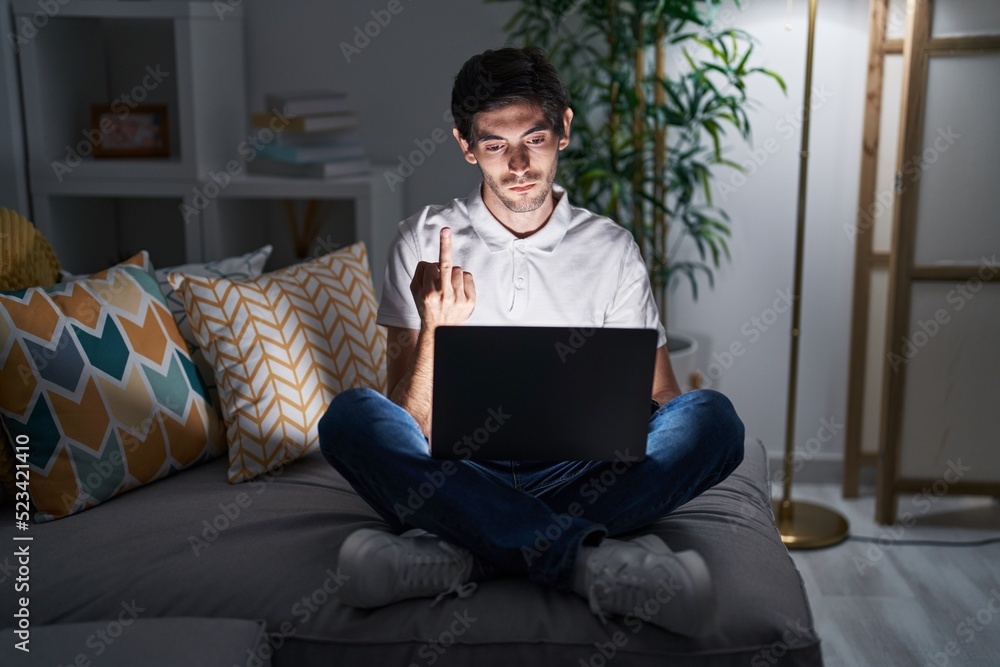 Young hispanic man using laptop at home at night showing middle finger, impolite and rude fuck off expression Young hispanic man using laptop at home at night showing middle finger, impolite and rude fuck off expression