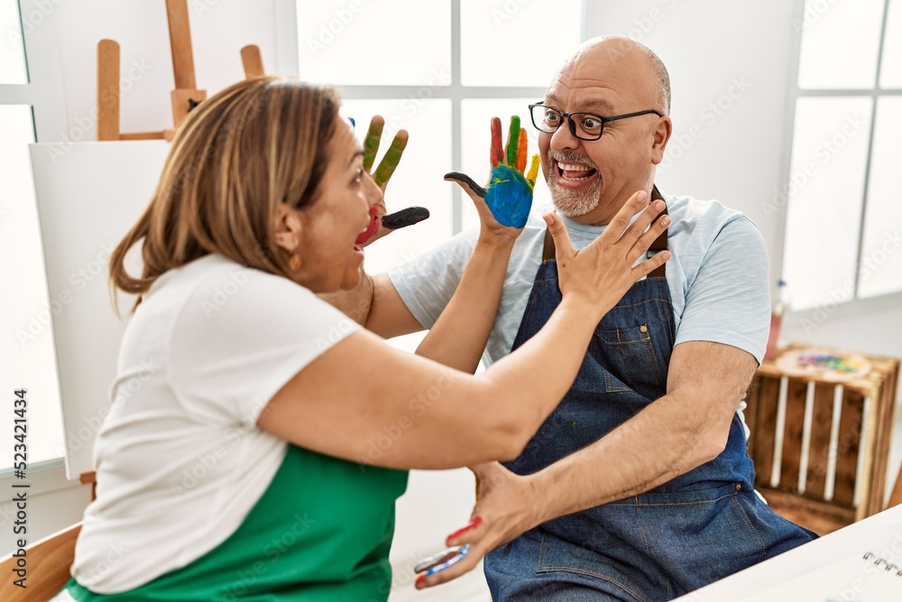Fototapeta premium Middle age hispanic painter couple smiling happy touching face with painted hands at art studio.