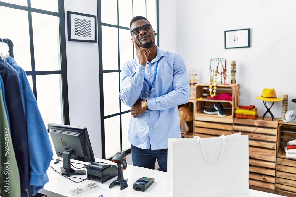 Young african man working as manager at retail boutique with hand on chin thinking about question, pensive expression. smiling with thoughtful face. doubt concept.