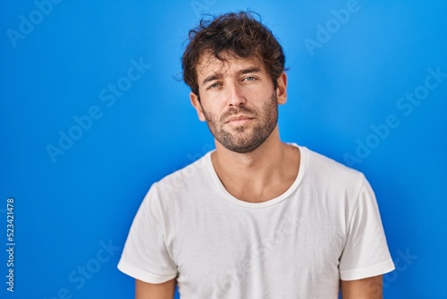 Hispanic young man standing over blue background looking sleepy and tired, exhausted for fatigue and hangover, lazy eyes in the morning.