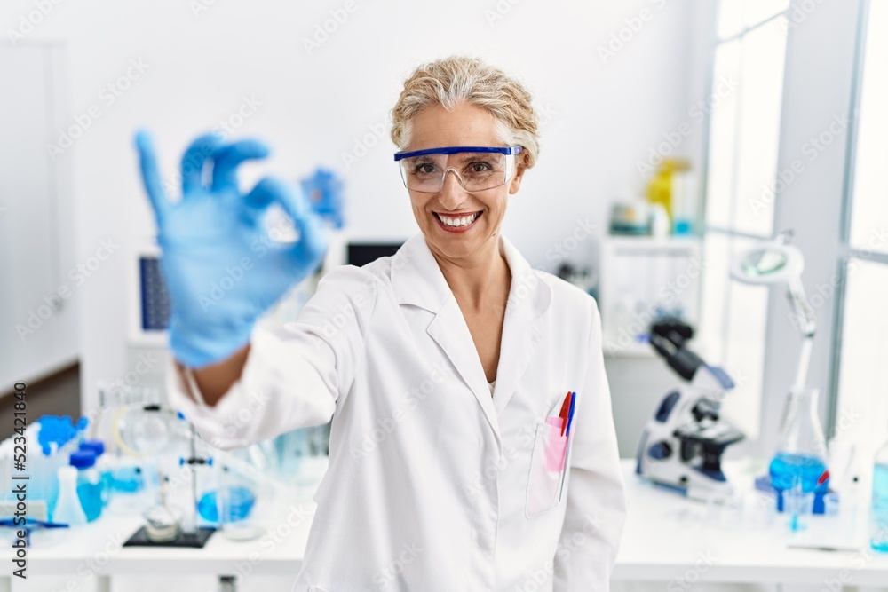 Middle age blonde woman working at scientist laboratory smiling positive doing ok sign with hand and fingers. successful expression.