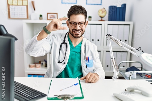 Young man with beard wearing doctor uniform and stethoscope at the clinic smiling and confident gesturing with hand doing small size sign with fingers looking and the camera. measure concept.