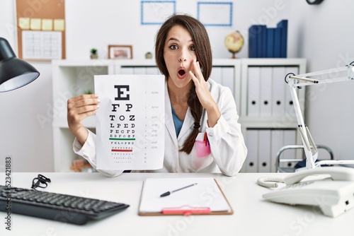 Young doctor woman holding eyesight test scared and amazed with open mouth for surprise, disbelief face