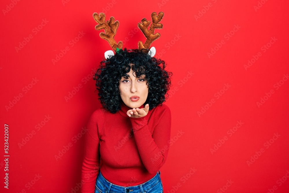 Young middle east woman wearing cute christmas reindeer horns looking at the camera blowing a kiss with hand on air being lovely and sexy. love expression.