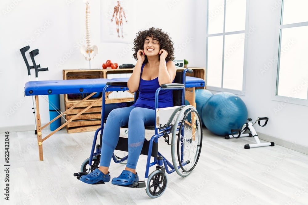 Obraz premium Young middle eastern woman sitting on wheelchair at physiotherapy clinic covering ears with fingers with annoyed expression for the noise of loud music. deaf concept.
