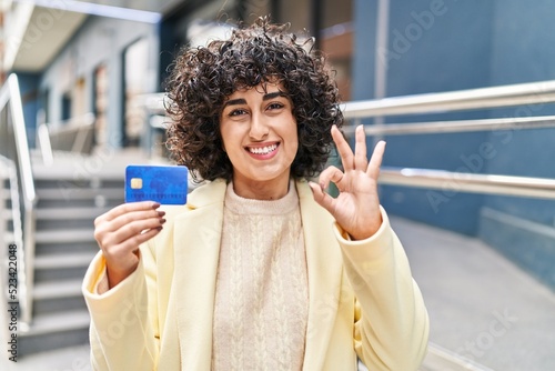Young brunette woman with curly hair holding credit card doing ok sign with fingers, smiling friendly gesturing excellent symbol