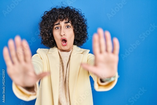Young brunette woman with curly hair standing over blue background doing stop gesture with hands palms, angry and frustration expression