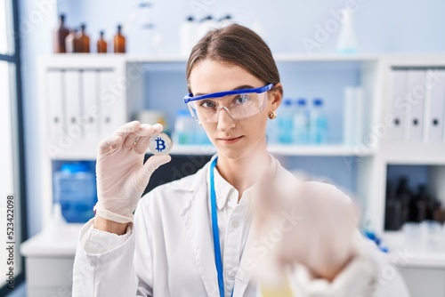 Young caucasian woman working at scientist laboratory holding bitcoin pointing with finger to the camera and to you, confident gesture looking serious