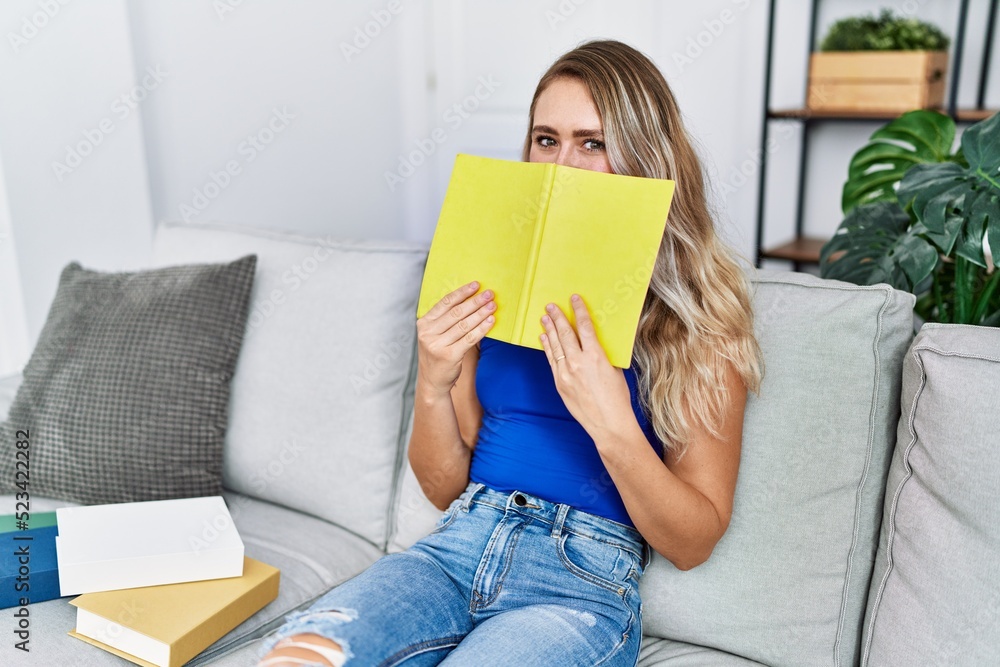 Young woman sitting on sofa covering face with book at home Young woman sitting on sofa covering face with book at home