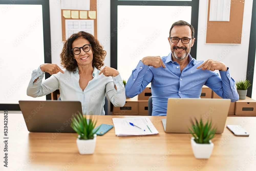 Middle age hispanic woman and man sitting with laptop at the office looking confident with smile on face, pointing oneself with fingers proud and happy. Middle age hispanic woman and man sitting with laptop at the office looking confident with smile on face, pointing oneself with fingers proud and happy.