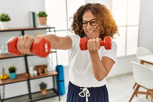 Middle age hispanic woman smiling confident training using dumbbells at home