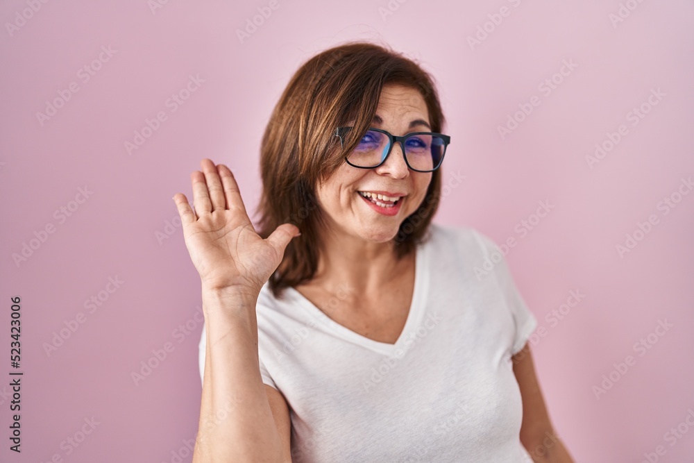 Middle age hispanic woman standing over pink background waiving saying hello happy and smiling, friendly welcome gesture
