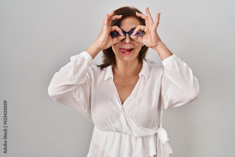 Middle age hispanic woman standing over isolated background doing ok gesture like binoculars sticking tongue out, eyes looking through fingers. crazy expression.