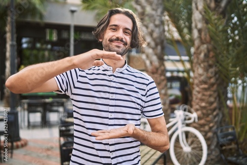 Young hispanic man with beard outdoors at the city gesturing with hands showing big and large size sign, measure symbol. smiling looking at the camera. measuring concept. © Krakenimages.com