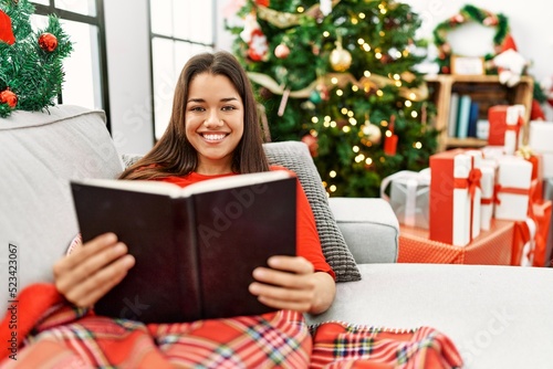 Young latin woman reading book sitting by christmas tree at home © Krakenimages.com