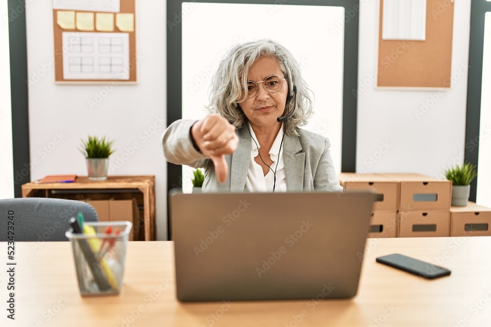Middle age businesswoman sitting on desk working using laptop at office looking unhappy and angry showing rejection and negative with thumbs down gesture. bad expression. Middle age businesswoman sitting on desk working using laptop at office looking unhappy and angry showing rejection and negative with thumbs down gesture. bad expression.