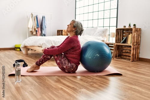 Middle age grey-haired woman training with fit ball sitting on the floor at bedroom.