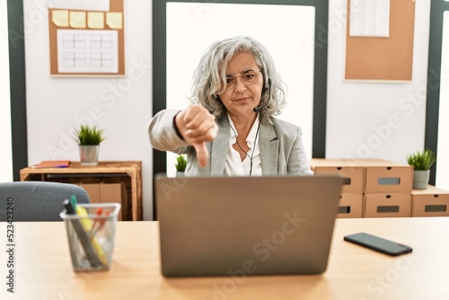 Middle age businesswoman sitting on desk working using laptop at office looking unhappy and angry showing rejection and negative with thumbs down gesture. bad expression.