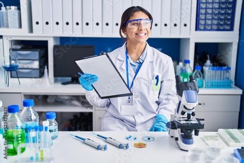 Hispanic young woman working at scientist laboratory looking away to side with smile on face  natural expression. laughing confident.