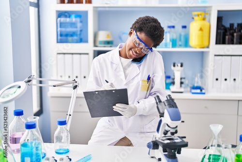 African american woman wearing scientist uniform talking on the smartphone write on clipboard at laboratory