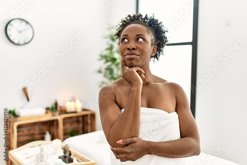 Young african american woman wearing towel standing at beauty center serious face thinking about question with hand on chin, thoughtful about confusing idea