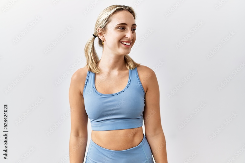 Young blonde woman wearing sportswear over isolated background looking away to side with smile on face, natural expression. laughing confident. Young blonde woman wearing sportswear over isolated background looking away to side with smile on face, natural expression. laughing confident.