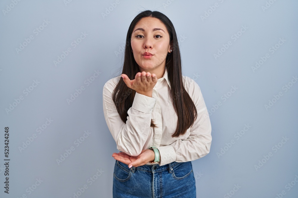 Young latin woman standing over blue background looking at the camera blowing a kiss with hand on air being lovely and sexy. love expression. Young latin woman standing over blue background looking at the camera blowing a kiss with hand on air being lovely and sexy. love expression.