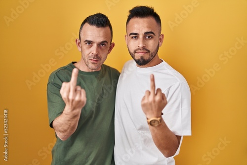 Homosexual couple standing over yellow background showing middle finger, impolite and rude fuck off expression