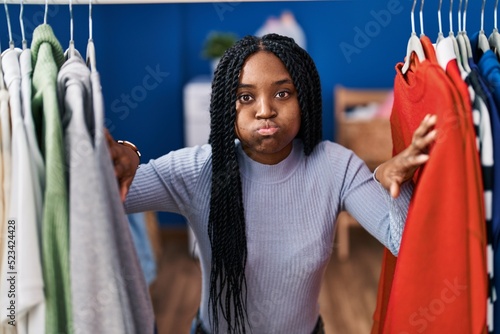 African american woman searching clothes on clothing rack puffing cheeks with funny face. mouth inflated with air, catching air.