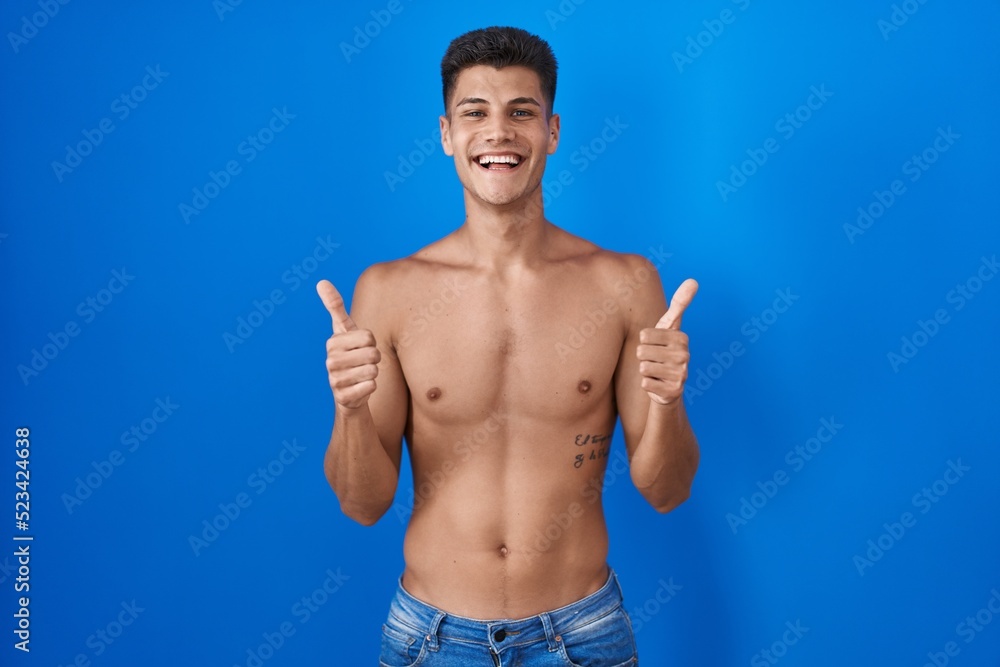 Young hispanic man standing shirtless over blue background success sign doing positive gesture with hand, thumbs up smiling and happy. cheerful expression and winner gesture.