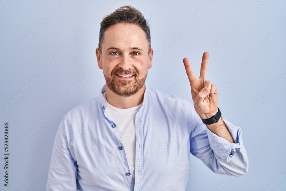 Middle age caucasian man standing over blue background showing and pointing up with fingers number two while smiling confident and happy. Middle age caucasian man standing over blue background showing and pointing up with fingers number two while smiling confident and happy.